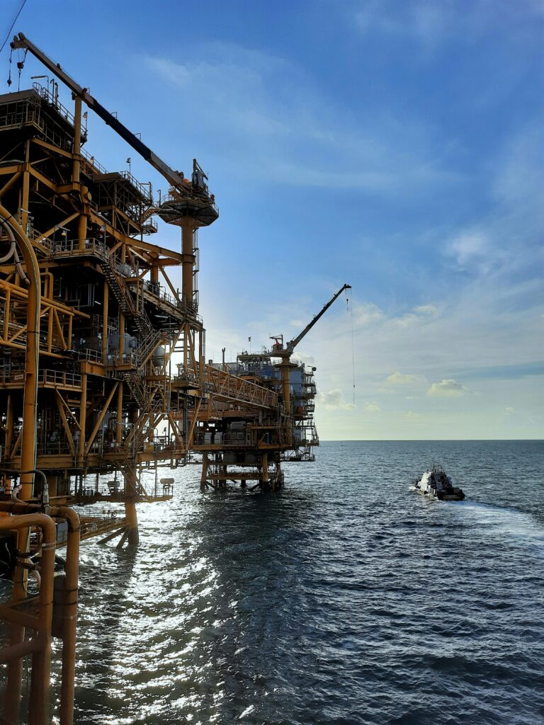 A large offshore oil rig with a supply vessel in the open sea under a blue sky and sunlight.